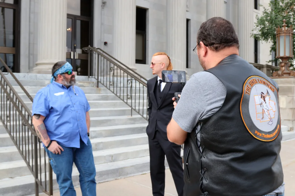 Steven Adelmund, Marketing Director for The Biker Lawyers, PC, stands with his back to the camera as he films Jon Jansen and a personal injury client in front of a courthouse in Cedar Rapids, Iowa.