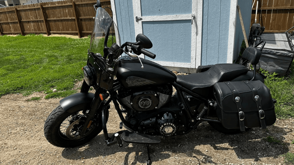 A Smoke black colored 2022 Indian Bobber Chief Dark Horse, parked on a gravel drive in front of a light blue shed.