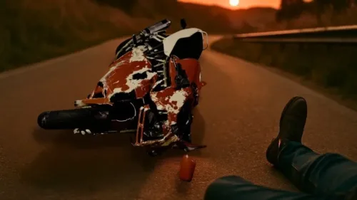 A badly damaged sport bike lays scraped and twisted on its side near a curve at sunset, with a booted leg visible, suggesting the rider is down off-frame.