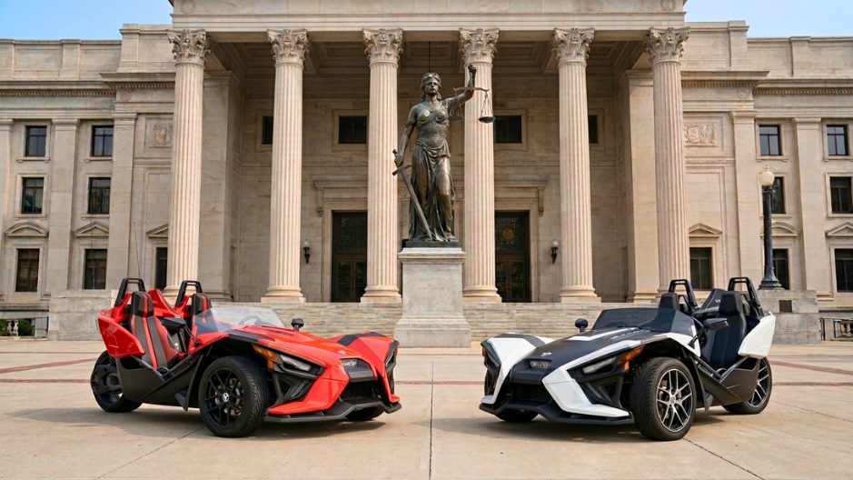 Two Polaris Slingshot autocycles parked in front of a courthouse with a Lady Justice statue and tall stone columns in the background.